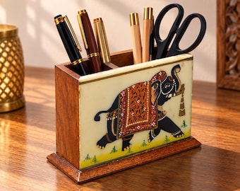 May include: A rectangular wooden desk organizer featuring a cream-colored front panel with a painted black elephant adorned in a red and orange patterned blanket. The organizer holds three pens and a pair of black scissors in its open compartments.
