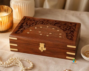 May include: A rectangular wooden box featuring intricate floral carvings on the lid, inlaid brass leaf patterns, and brass corner accents. The box includes a central brass latch closure on the front, showcasing a dark brown wood grain finish.