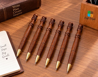 May include: Six ballpoint pens with dark brown wood barrels and gold-toned metal tips. Each pen features a decorative turned wood design at the top. The pens are arranged in a row on a wooden surface next to a notebook with the text Small Steps Big Goals.