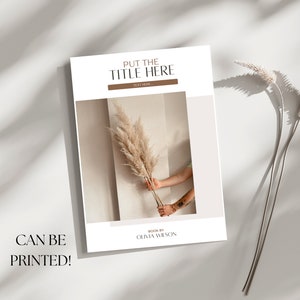 May include: A white book cover with the text "PUT THE TITLE HERE" and "TEXT HERE" in brown text. The cover also features a photo of a hand holding a dried pampas grass against a beige wall. The text "BOOK BY OLIVIA WILSON" is at the bottom of the cover.