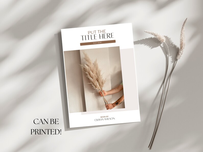 May include: A white book cover with the text "PUT THE TITLE HERE" and "TEXT HERE" in brown text. The cover also features a photo of a hand holding a dried pampas grass against a beige wall. The text "BOOK BY OLIVIA WILSON" is at the bottom of the cover.