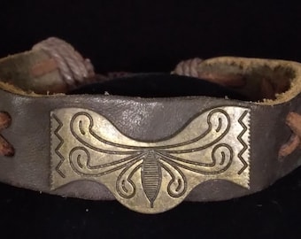 May include: A dark brown leather bracelet featuring a central rectangular brass plate with an engraved symmetrical floral-like pattern and zigzag borders. The leather band is secured with brown cord lacing on both sides of the metal centerpiece.
