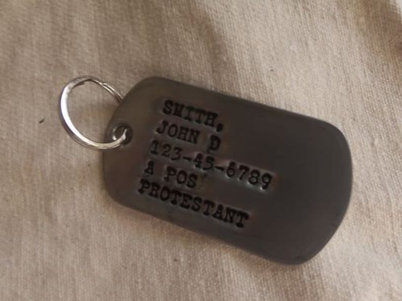 K&ouml;nnte beinhalten: Ein silberner Metall-Hundeanh&auml;nger mit dem Text "SMITH, JOHN D 123-45-6789 A POS PROTESTANT" eingraviert.