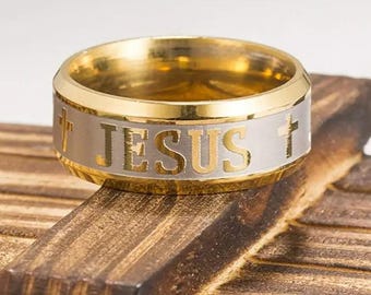 May include: A gold and silver-toned band ring featuring the word JESUS engraved in gold lettering on a silver center band, flanked by two gold crosses. The ring has polished gold edges and a wide, flat profile with a metallic finish throughout.