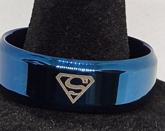 May include: A polished blue band ring featuring a silver-toned Superman logo engraved on the front surface. The ring has a smooth, reflective finish and a beveled edge design, showcasing the iconic diamond-shaped emblem with the letter S inside.