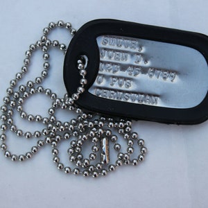 May include: A silver dog tag with a black rubber cover and a silver chain. The tag has the text "SMITH, JOHN J. 125-45-6789 0 POS  CERKSUWAN" engraved on it.