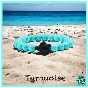 Op de afbeelding: Turquoise kralen armband met een zwarte schildpad bedel. Het armband ligt op een zandstrand met een blauwe oceaan op de achtergrond.