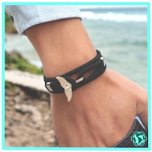 May include: A black braided bracelet with a silver whale tail charm. The bracelet is worn on a person's wrist.