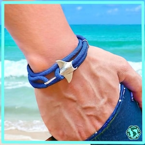 Op de afbeelding: Een blauwe touw armband met een zilveren mantarog charme. De armband wordt gedragen op iemands pols.