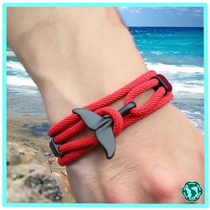 May include: Red braided rope bracelet with a black whale tail charm. The bracelet is worn on a person's wrist.