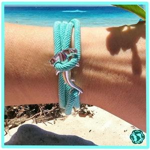 May include: A turquoise rope bracelet with a silver hammerhead shark charm. The bracelet is worn on a person's wrist against a beach background.