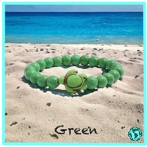 Könnte beinhalten: Ein grünes Perlenarmband mit einem schildkrötenförmigen Verschluss liegt auf einem Sandstrand. Das Armband besteht aus runden, hellgrünen Perlen. Das Wort "Green" steht in schwarzer Schrift darunter, mit einem türkisfarbenen Rand.