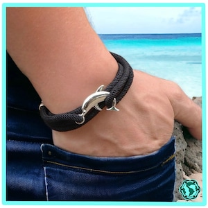 Op de afbeelding: Een zwarte gevlochten armband met een zilveren dolfijnbedel. De armband wordt gedragen op de pols van een persoon tegen een achtergrond van een strand en oceaan.
