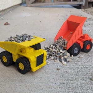 Könnte beinhalten: Ein leuchtend gelber Muldenkipper gefüllt mit kleinem grauem Kies und ein leuchtend orangefarbener Muldenkipper mit gekippter Ladefläche, der einen Haufen grauen Kies auf den Boden schüttet. Beide Spielzeugfahrzeuge haben schwarze Räder.