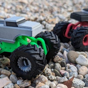 May include: Two toy monster trucks with oversized black tires. The front truck has a light gray body and a bright green chassis. The rear truck features a matte black body with a red chassis and red wheel rims, both showcasing detailed tread.