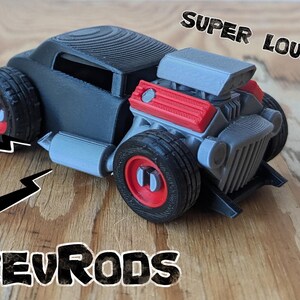 Può includere: Un'auto giocattolo stampata in 3D con carrozzeria nero opaco, blocco motore grigio e coperchi delle valvole rosso brillante. Le ruote presentano cerchi rossi e centri grigi. Il testo SUPER LOUD!! appare sopra e REVRODS è stampato sotto.