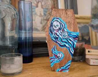May include: A wooden coffin-shaped box painted with blue, white, and red swirls and three white skulls.