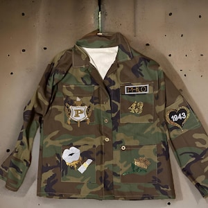 Op de afbeelding: Een camouflagejas met verschillende patches, waaronder een gouden "PHILO"-patch, een gouden "43"-patch, een gouden "P"-patch, een gouden hart met "1943" erin, een gouden roos en een gouden lippenontwerp.