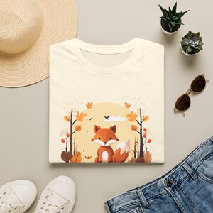 Könnte beinhalten: Ein cremefarbenes T-Shirt mit einer grafischen Illustration eines Fuchses, der in einer Waldumgebung mit Herbstblättern, kahlen Bäumen und einer großen gelben Sonne im Hintergrund sitzt. Das Motiv ist mittig auf der Brust platziert.