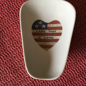 May include: White ceramic spoon rest shaped like a heart with a distressed American flag design and the text "Land That I Love".