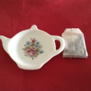 May include: A white porcelain teapot-shaped tea bag holder with a floral design in pink and blue. A tea bag rests beside the holder on a red surface. The holder has a handle and spout.