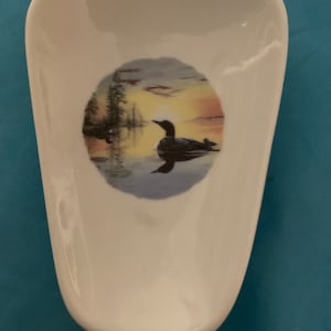 May include: White ceramic spoon rest with a colorful image of a loon bird in a lake at sunset.