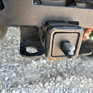 May include: A black metal trailer hitch receiver with a silver ball hitch pin. The hitch is mounted on a vehicle.