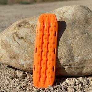 Könnte beinhalten: Orangefarbene Traktionsmatte mit einer strukturierten Oberfläche und mehreren erhabenen Noppen. Die Matte ist so konzipiert, dass sie Fahrzeugen in rutschigen Bedingungen mehr Grip verschafft.