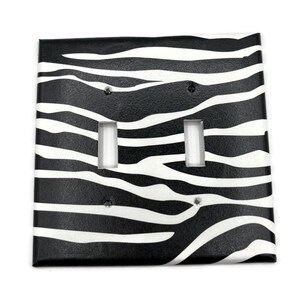 May include: A square double light switch wall plate featuring a black and white zebra stripe pattern. The cover has two rectangular cutouts for toggle switches and four small circular holes for mounting screws, arranged in a standard configuration.