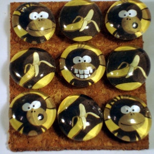 May include: Nine round glass monkey push pins with a yellow background. Each pin features a different monkey design with a banana. Some monkeys are smiling, some are holding bananas, and some are wearing hats.