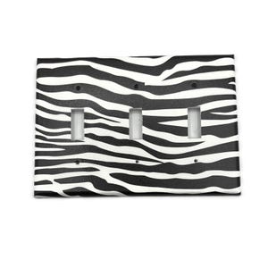 May include: A rectangular triple toggle light switch wall plate featuring a black and white zebra stripe pattern. The cover includes three vertical rectangular cutouts for switches and six small circular holes for mounting screws to attach it.