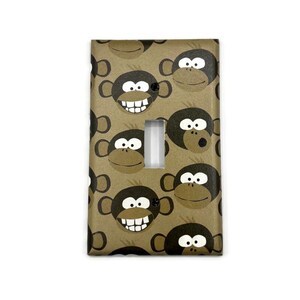 May include: A rectangular light switch cover plate featuring a repeating pattern of cartoon monkey faces on a tan background. The monkeys have dark brown ears and facial features, white eyes, and some display wide grins with visible white teeth.