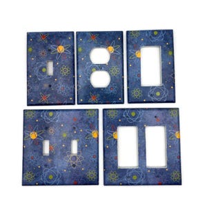 May include: Set of five blue light switch and outlet covers with a retro atomic pattern. The covers feature a dark blue background with yellow, green, and white atomic symbols.