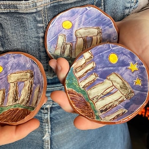 May include: Three hand-painted ceramic dishes with a purple background and a depiction of Stonehenge. The dishes have a brown rim and a yellow sun or stars.