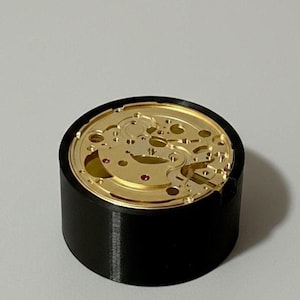 May include: A detailed view of a watch mechanism, featuring gold-coloured gears and tiny parts. The mechanism is housed in a black cylindrical casing, providing a strong contrast. The design highlights the precision and craftsmanship of watchmaking.