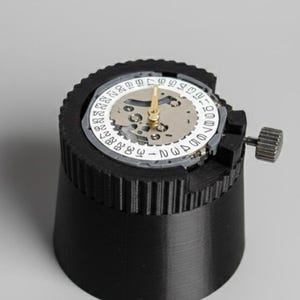 Könnte beinhalten: Ein schwarzer Uhrenwerkhalter mit eingebautem Uhrwerk. Das Uhrwerk hat ein weißes Zifferblatt mit schwarzen Zahlen und goldenen Zeigern. Der Halter hat ein geripptes Design und einen kleinen Einstellknopf.