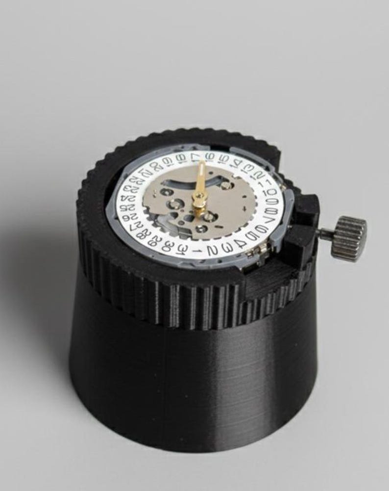 May include: A black watch movement holder with a watch movement installed. The watch movement has a white dial with black numbers and gold hands. The holder has a ribbed design and a small adjustment knob.