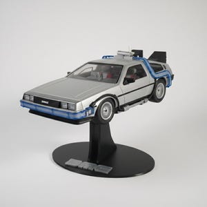 Può includere: Modello argentato della DeLorean, con dettagli blu e neri, montato su un supporto nero. L'auto presenta elementi di design dettagliati ed è un oggetto da collezione. Il supporto ha le lettere DMC.