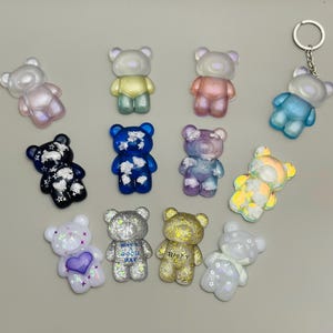 May include: A collection of colorful bear-shaped charms. The charms are in various colors, including blue, pink, yellow, and iridescent. Some charms have glitter or text that says "Have a good day" and "Happy". One charm is attached to a keychain.