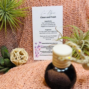 May include: A small glass bottle with a black cap and a white label that reads "Clean and Fresh" with the ingredients and fragrance listed. The bottle is sitting on a pink and white fuzzy surface.