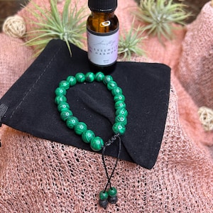 May include: A green malachite beaded bracelet with black string ties and a small bottle of essential oil labeled "Essential Harmony" on a black velvet pouch.