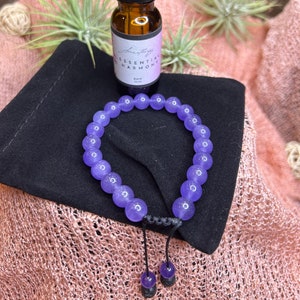 May include: A purple amethyst bracelet with a black cord and a small bottle of essential oil labeled "Essential Harmony" on a pink and black fabric background.