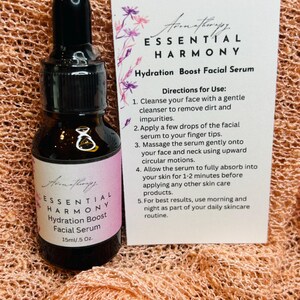 May include: Aromatherapy Essential Harmony Hydration Boost Facial Serum in a dark glass bottle with a dropper. The label reads 15ml/.5 oz. A card provides directions for use. The background is a peach-colored fabric.