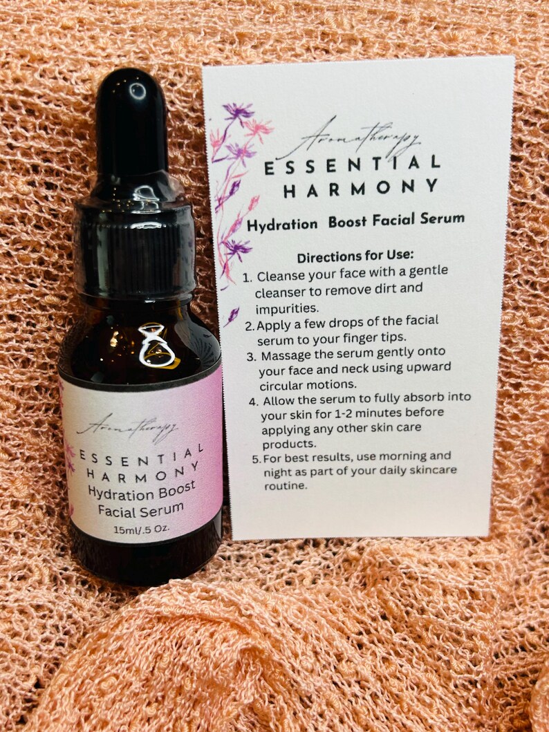 May include: Aromatherapy Essential Harmony Hydration Boost Facial Serum in a dark glass bottle with a dropper. The label reads 15ml/.5 oz. A card provides directions for use. The background is a peach-colored fabric.