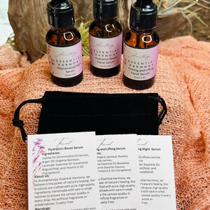 May include: Three small amber glass bottles of facial serum with black droppers, labeled "Essential Harmony." The bottles are on a peach-colored surface with product information cards and a black pouch. The labels have pink accents.