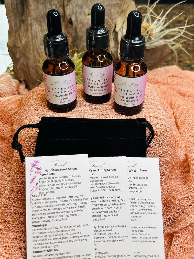May include: Three small amber glass bottles of facial serum with black droppers, labeled "Essential Harmony." The bottles are on a peach-colored surface with product information cards and a black pouch. The labels have pink accents.