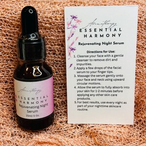 May include: A dark glass bottle of Essential Harmony Rejuvenating Night Serum, 15ml/.5 Oz, with a black dropper. A white card with instructions and the product name is next to the bottle. The background is a peach-colored, textured fabric.