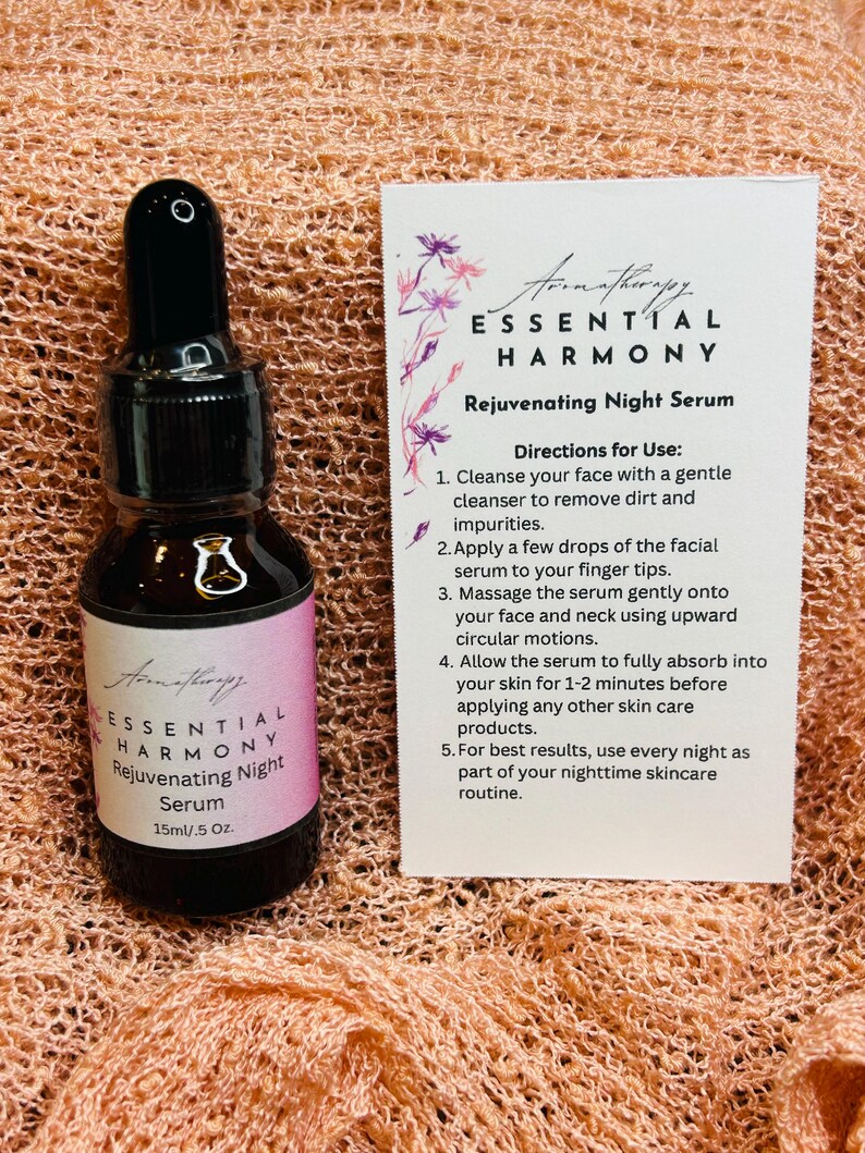 May include: A dark glass bottle of Essential Harmony Rejuvenating Night Serum, 15ml/.5 Oz, with a black dropper. A white card with instructions and the product name is next to the bottle. The background is a peach-colored, textured fabric.