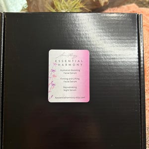 May include: A black box with a pink and white label. The label reads "ESSENTIAL HARMONY" and lists facial serums: Hydration Boosting, Firming and Lifting, and Rejuvenating Night Serum. The website Aessentialharmony.etsy.com is also listed.
