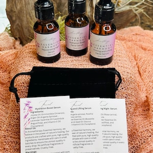 May include: Three small amber glass bottles of facial serum with black droppers and pink labels, labeled "Essential Harmony." The bottles are displayed with a black pouch and ingredient cards. The background is a light peach fabric.